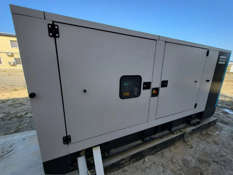 Atlas Copco generator in a sound-attenuated enclosure with a controller window in the door, installed on an open site next to a building.